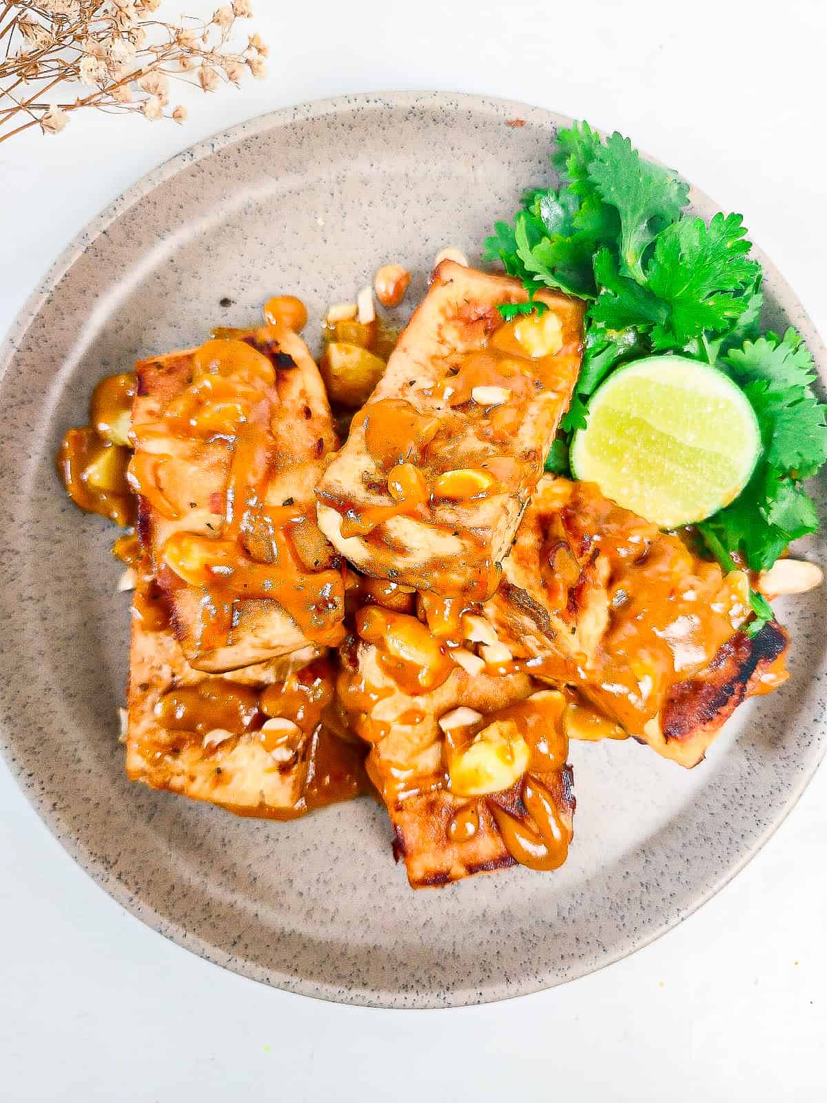 Crispy peanut satay tofu topped with creamy peanut sauce, chopped peanuts, fresh cilantro, and lime on a serving plate.