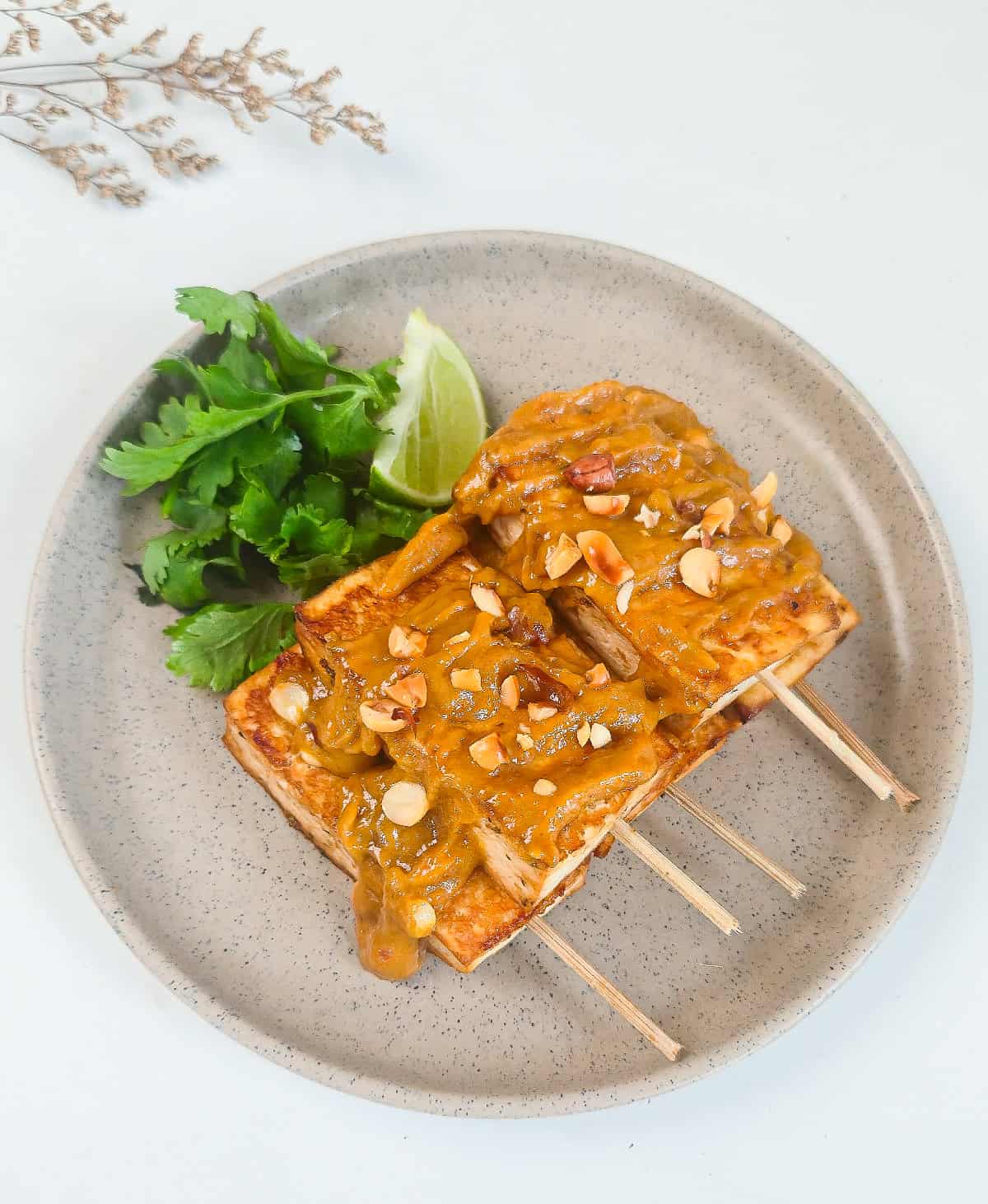 Peanut satay tofu skewers topped with creamy peanut sauce, chopped peanuts, cilantro, and lime on a ceramic plate.