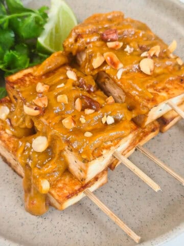 Close-up of crispy peanut satay tofu skewers covered in rich peanut sauce and chopped peanuts with fresh cilantro and lime.