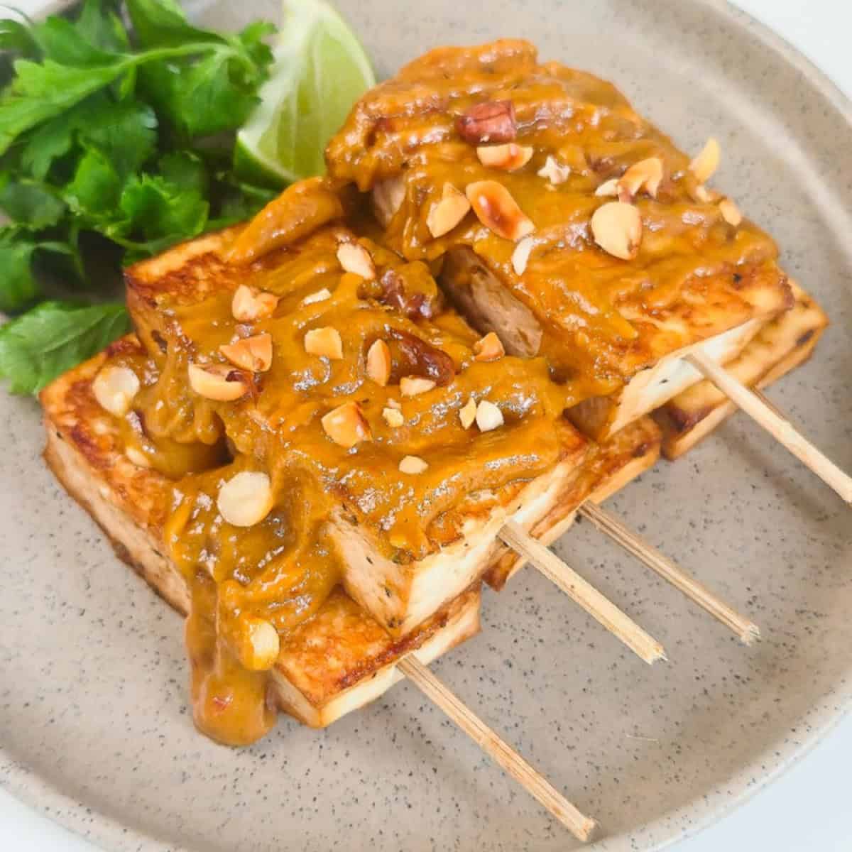 Close-up of crispy peanut satay tofu skewers covered in rich peanut sauce and chopped peanuts with fresh cilantro and lime.