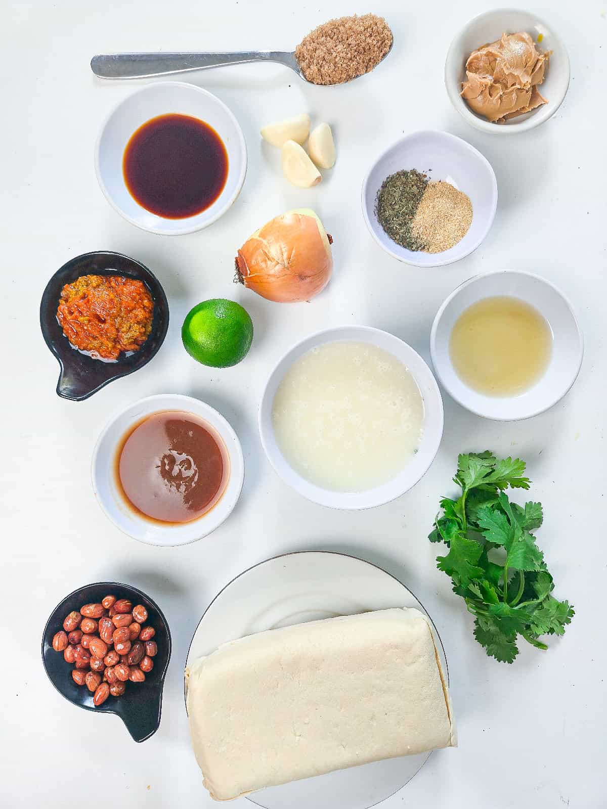 Ingredients for peanut satay tofu including tofu, peanut butter, soy sauce, coconut milk, onion, garlic, lime, curry paste, peanuts, and fresh cilantro arranged on a white background.