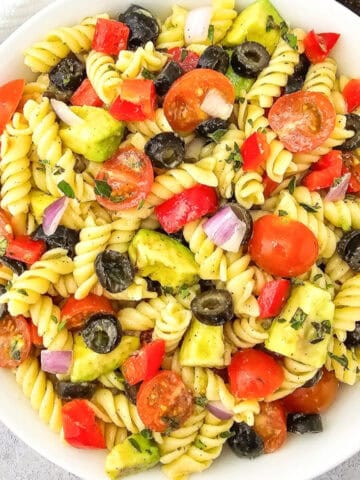 Overhead view of vegan pasta salad with rotini, avocado, cherry tomatoes, black olives, red bell pepper, and red onion in a white bowl.