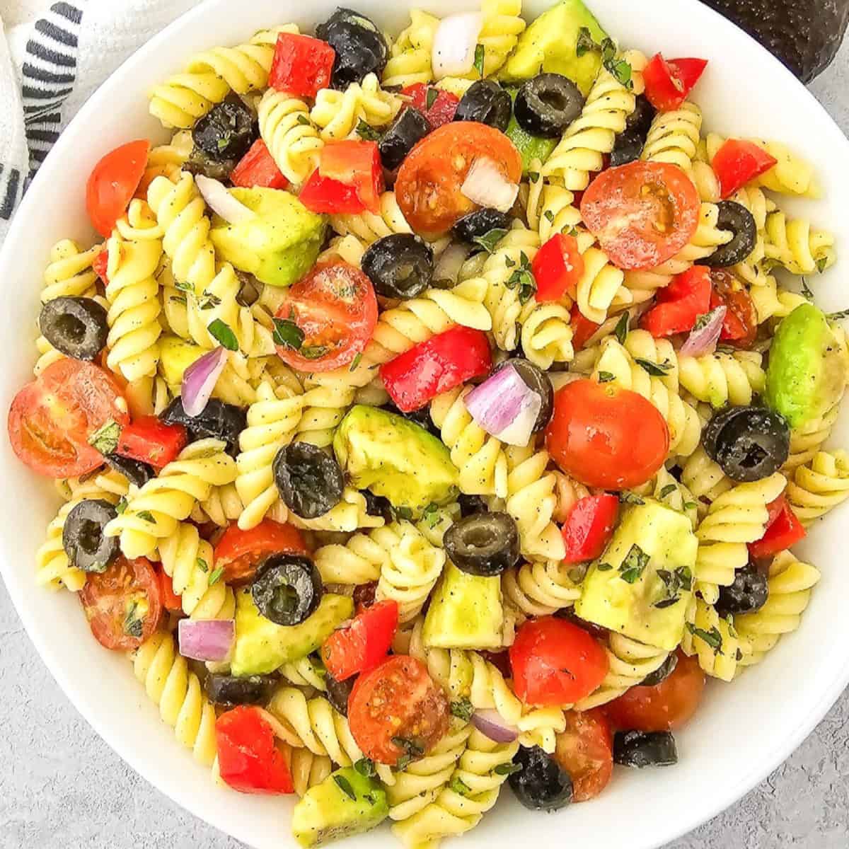 Overhead view of vegan pasta salad with rotini, avocado, cherry tomatoes, black olives, red bell pepper, and red onion in a white bowl.