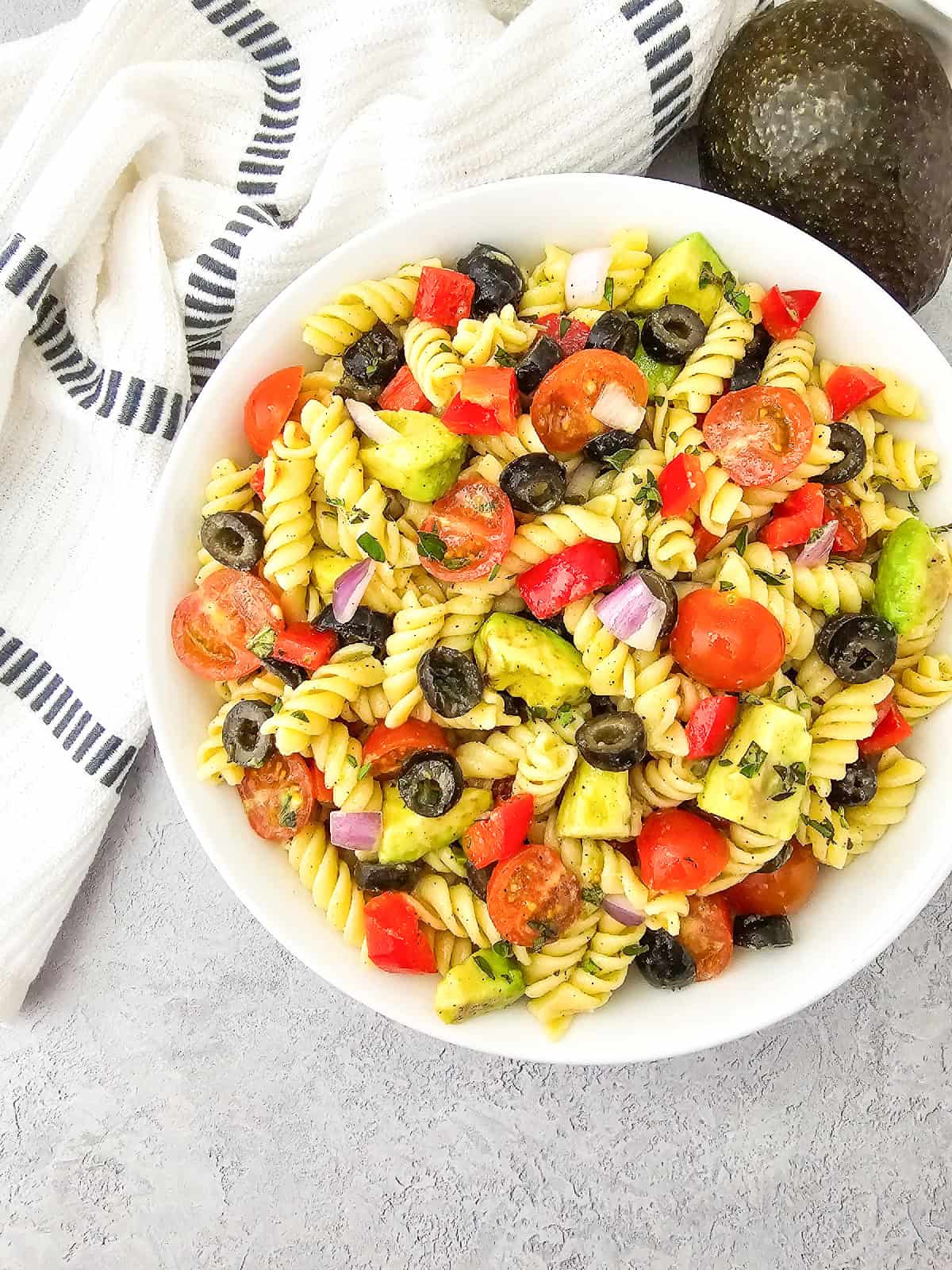 Overhead view of vegan pasta salad with rotini, avocado, cherry tomatoes, black olives, red bell pepper, and red onion in a white bowl.