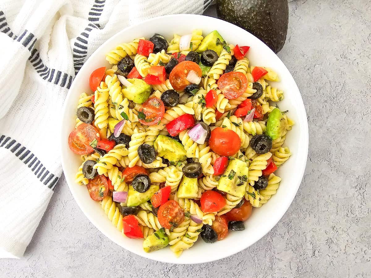 Vegan pasta salad served in a white bowl on a light gray surface with a striped kitchen towel and avocado in the background.