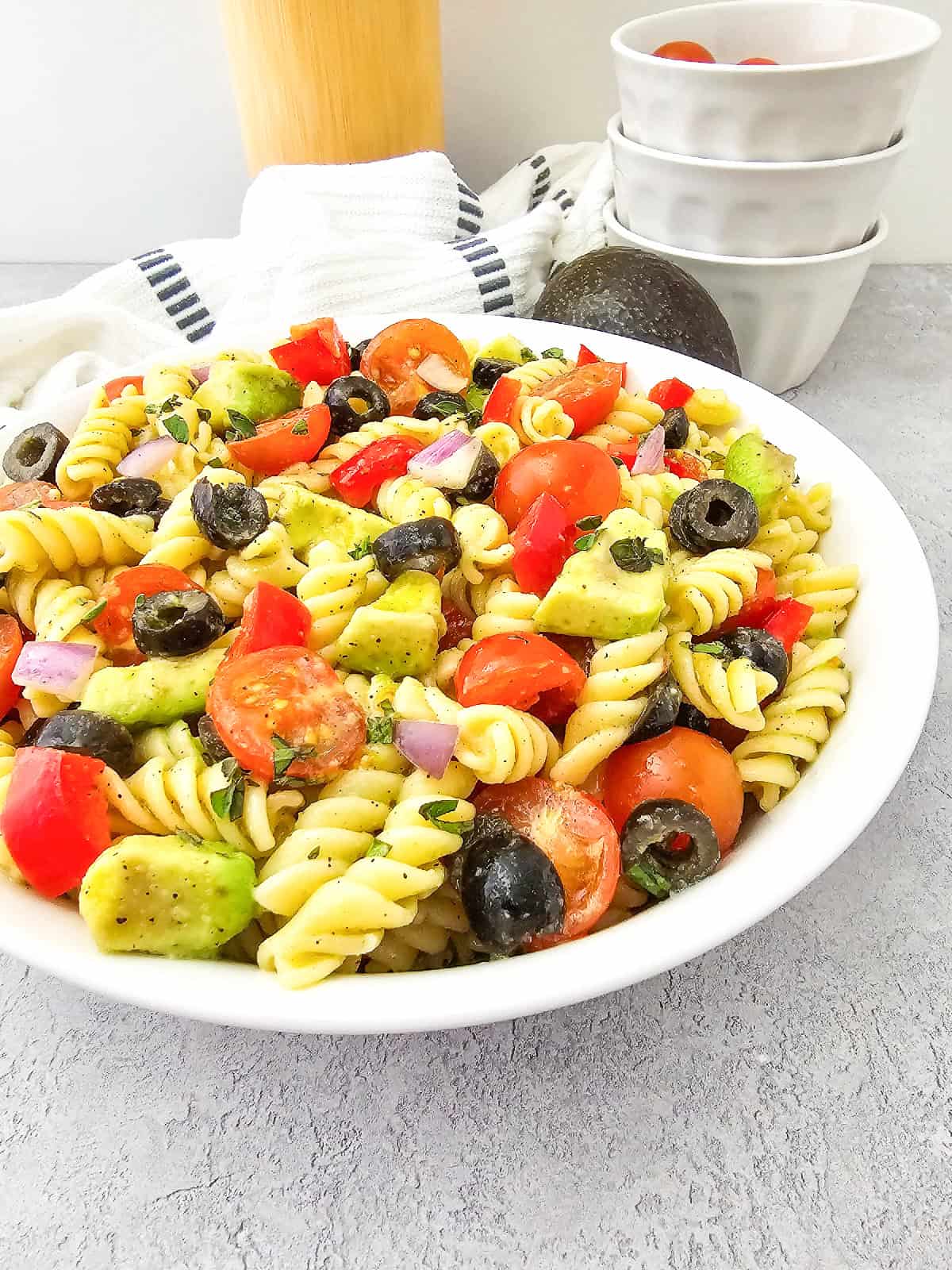 Side-angle view of vegan pasta salad with rotini, avocado, cherry tomatoes, black olives, and red bell pepper in a white serving bowl.