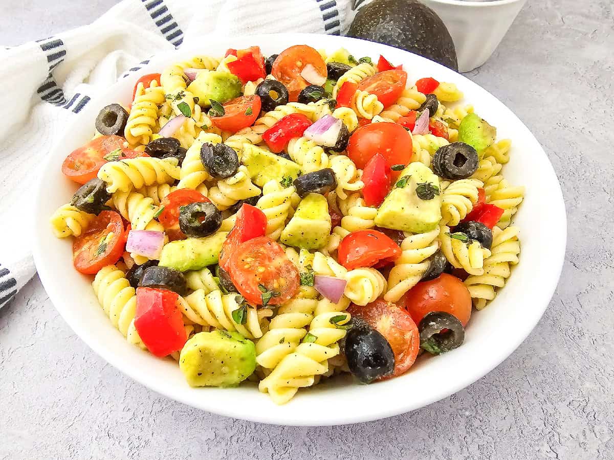 Angled view of vegan pasta salad with rotini pasta, avocado, cherry tomatoes, olives, and bell pepper in a white bowl.