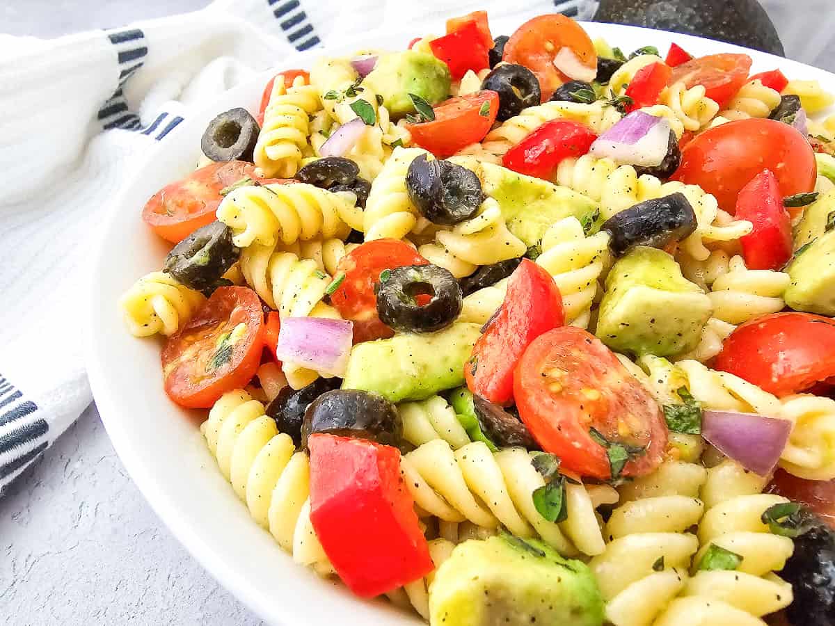 Extreme close-up of vegan pasta salad showing rotini pasta coated in dressing with avocado, cherry tomatoes, black olives, and red bell pepper.