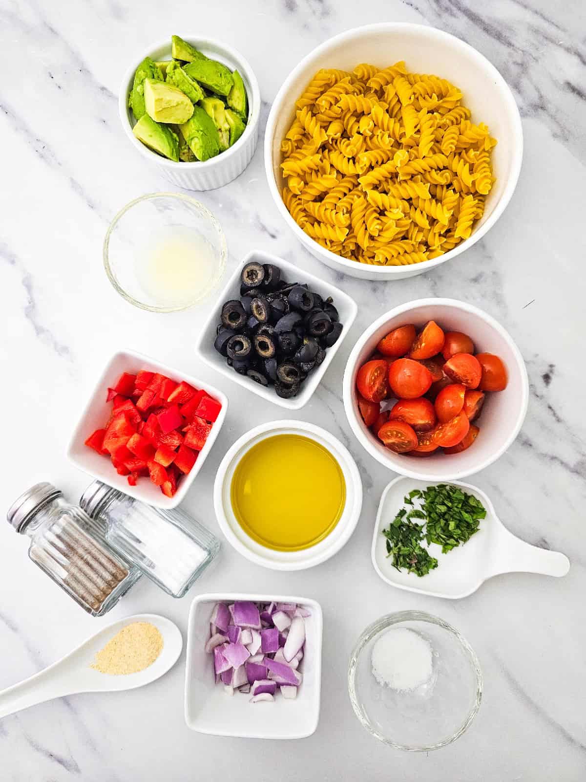 Ingredients for vegan pasta salad arranged on a marble countertop, including cooked rotini pasta, avocado, cherry tomatoes, black olives, red bell pepper, red onion, fresh basil, olive oil, lemon juice, and seasonings.