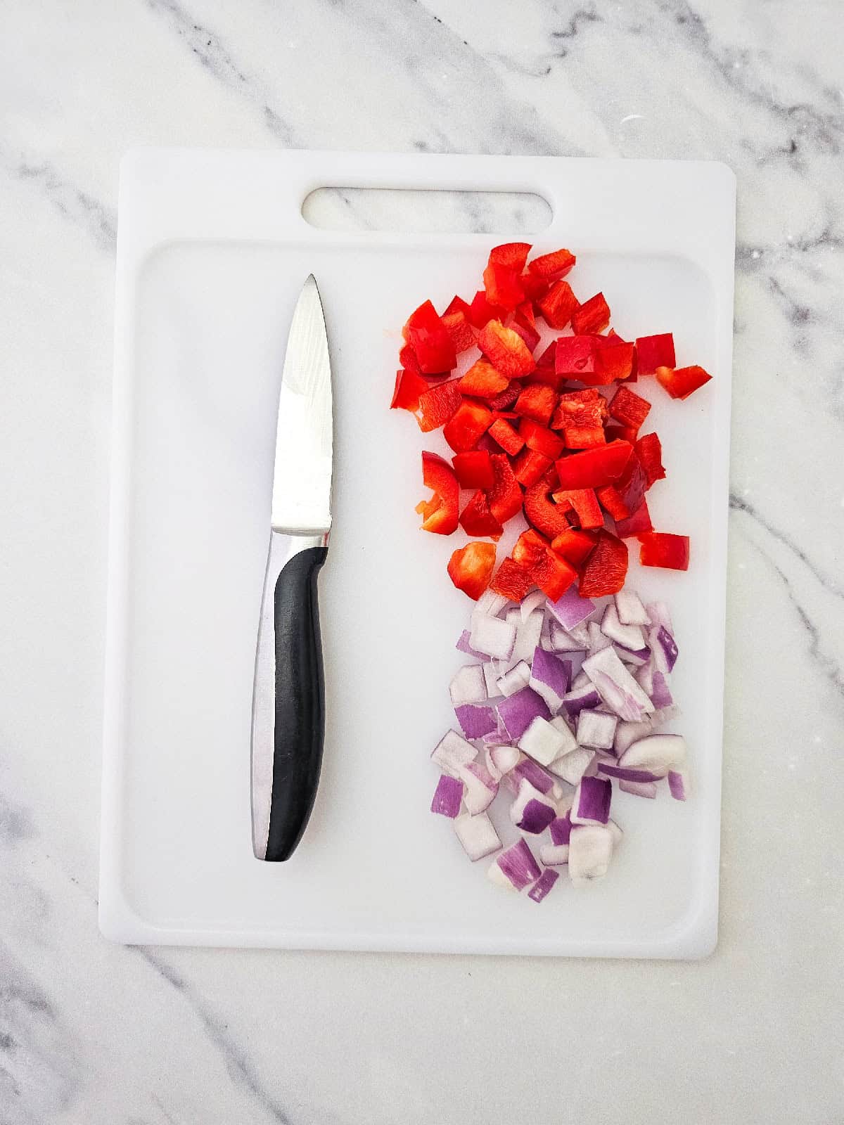 Chopped red bell pepper and diced red onion on a white cutting board with a kitchen knife.
