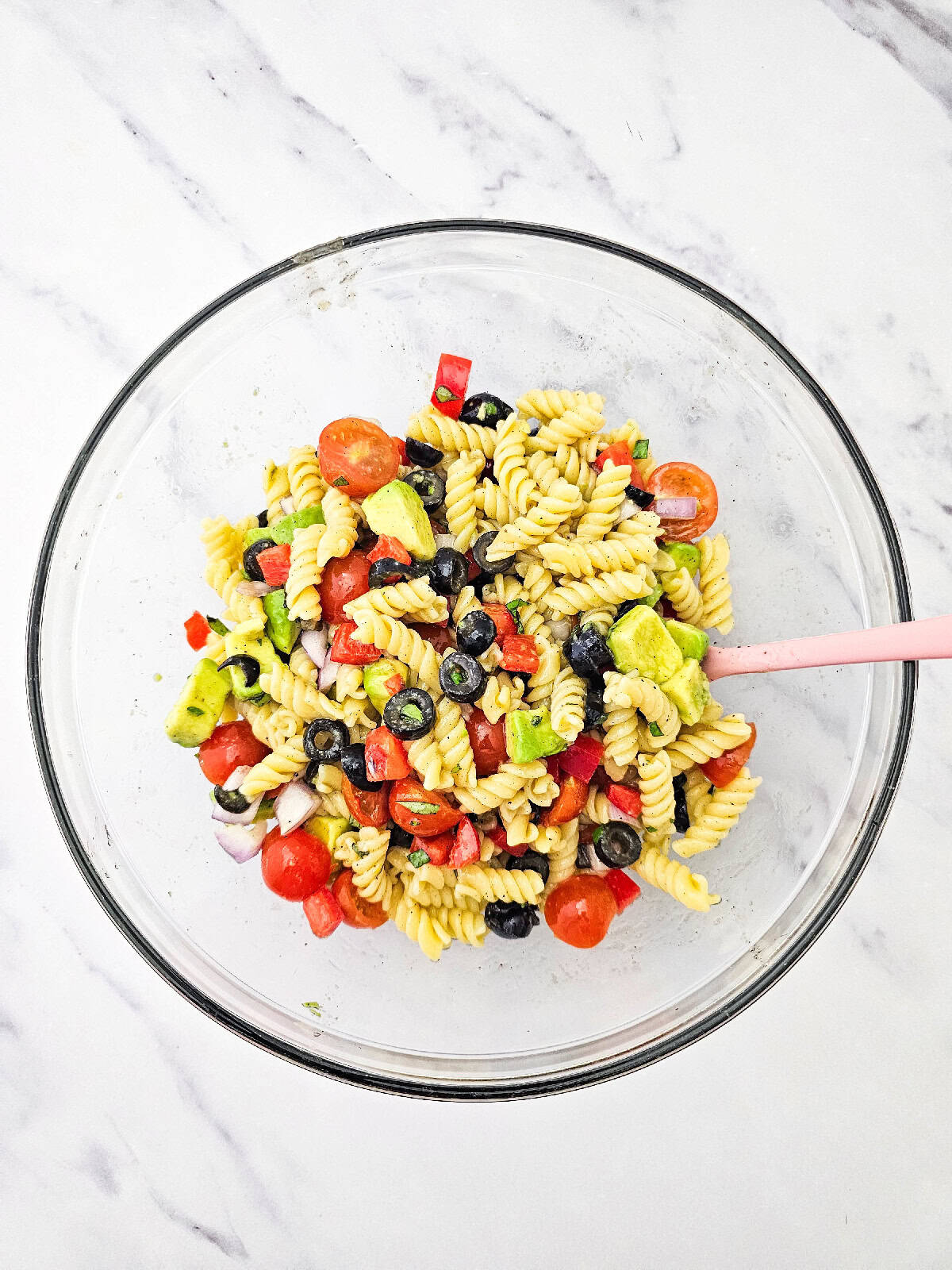 Vegan pasta salad tossed with avocado, cherry tomatoes, black olives, red bell pepper, red onion, basil, and lemon dressing in a mixing bowl.