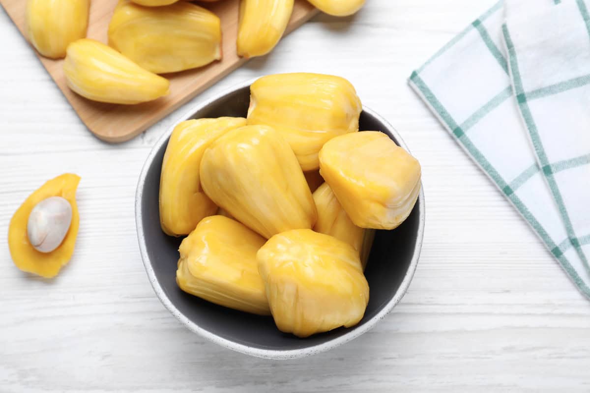 Sweet yellow jackfruit pods in a bowl on a white table with a jackfruit seed nearby.