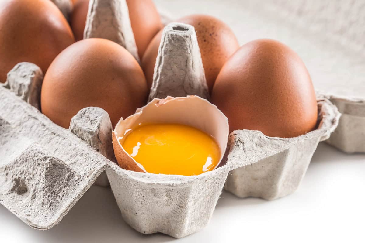 Brown eggs in a carton with a cracked egg shell holding a fresh raw yolk.