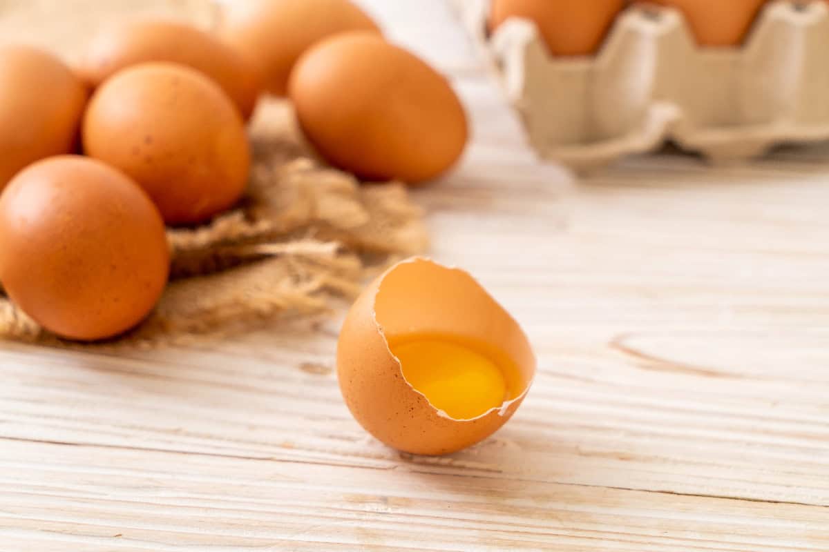 Cracked brown egg shell containing raw yolk on a wooden table surrounded by fresh eggs.