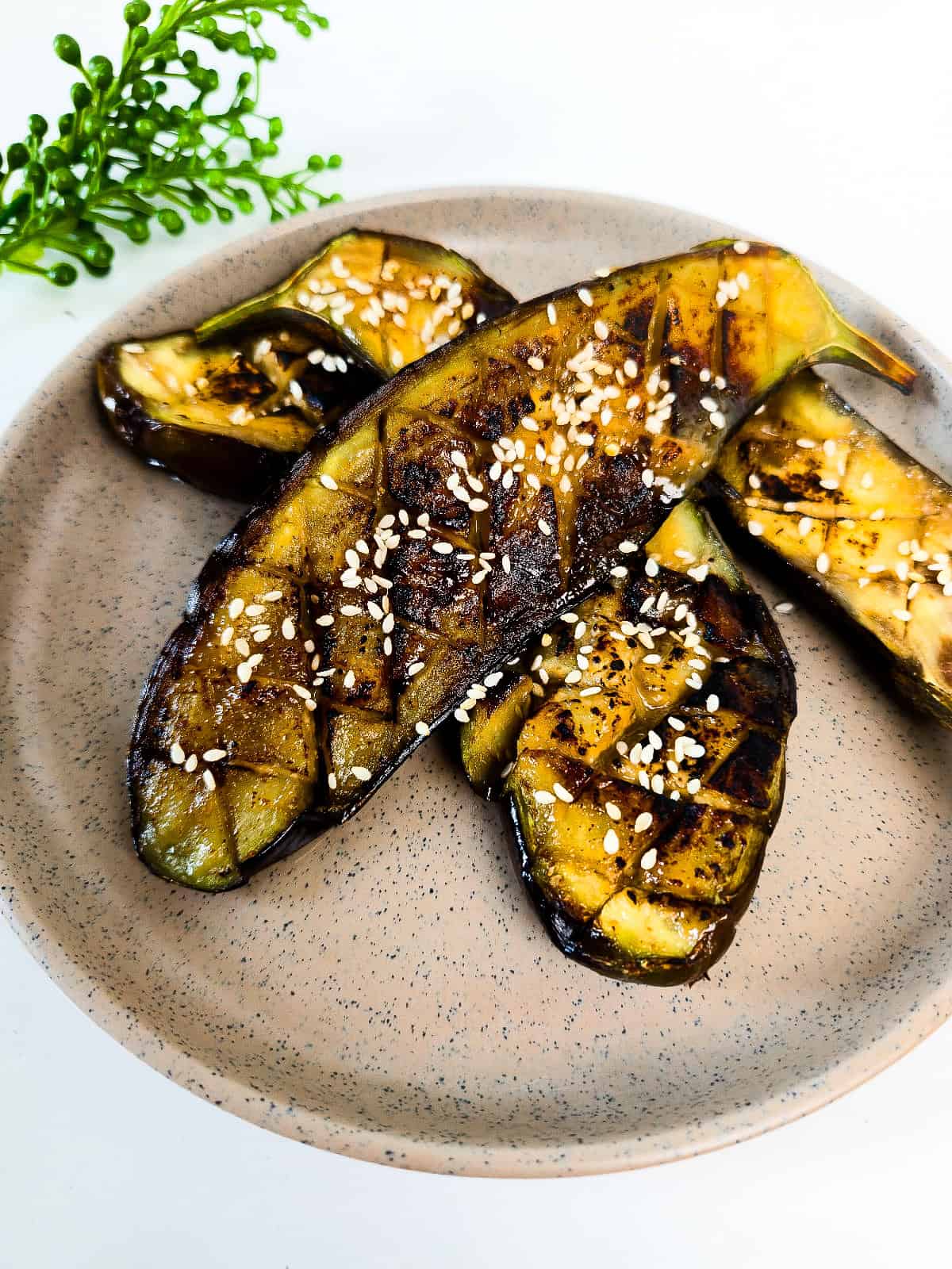 Roasted miso glazed eggplant halves topped with sesame seeds and served on a speckled ceramic plate.