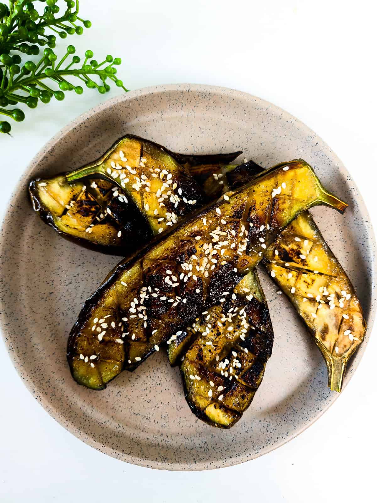 Top-down view of roasted miso glazed eggplant halves topped with sesame seeds and arranged on a speckled ceramic plate.