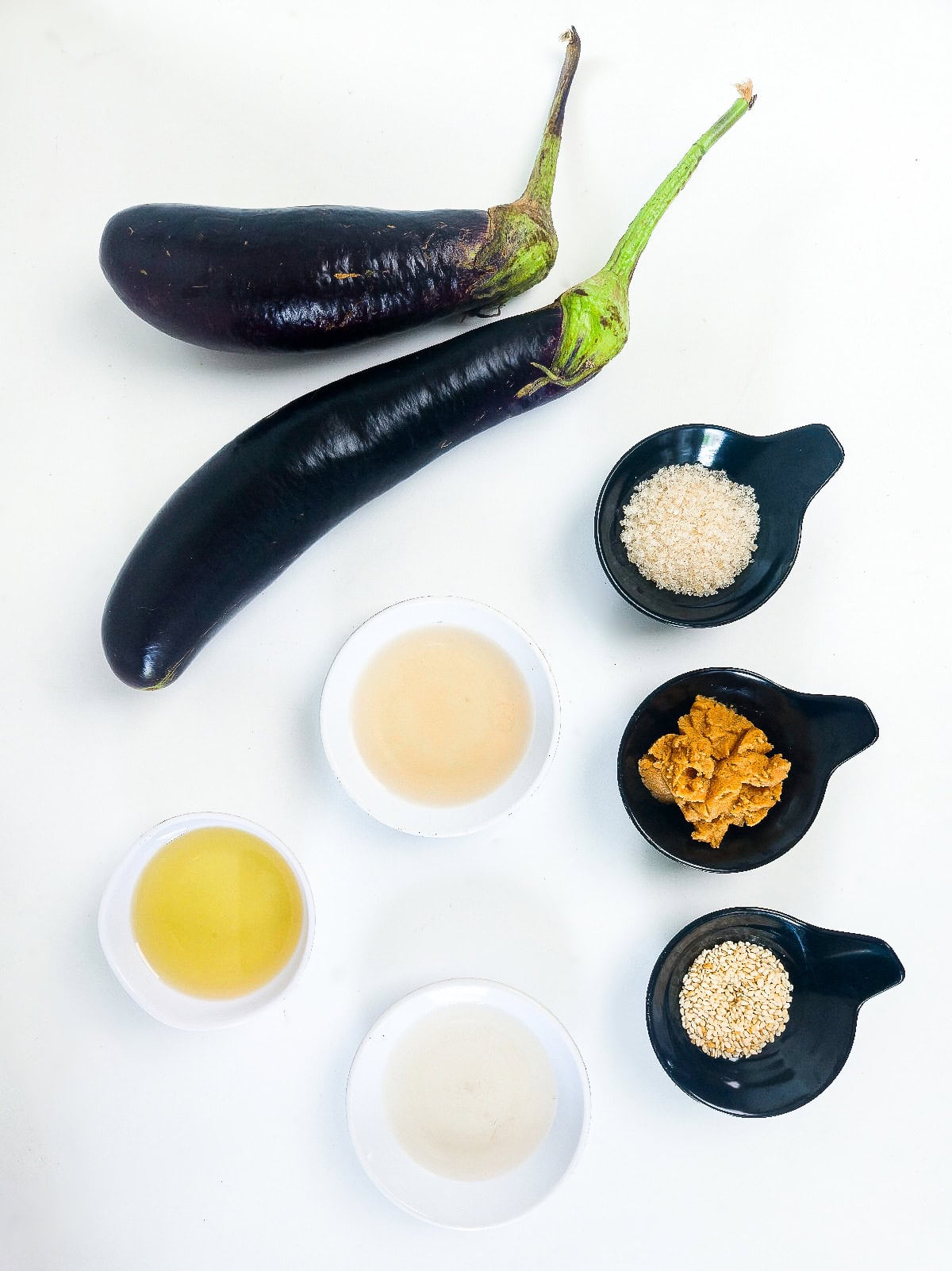 Ingredients for miso glazed eggplant including Japanese eggplants, miso paste, mirin, sesame seeds, oil, and sugar arranged on a white background.