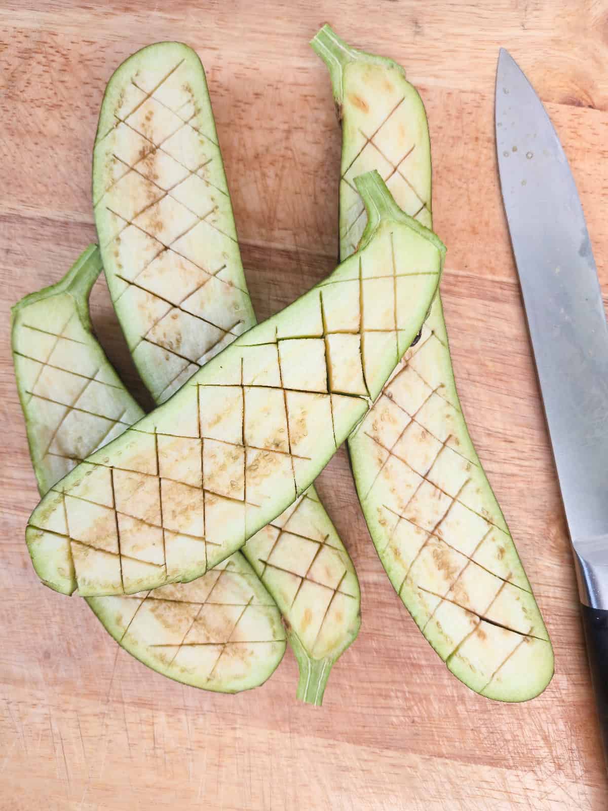 Japanese eggplants scored in a crosshatch pattern on a wooden cutting board next to a chef's knife.