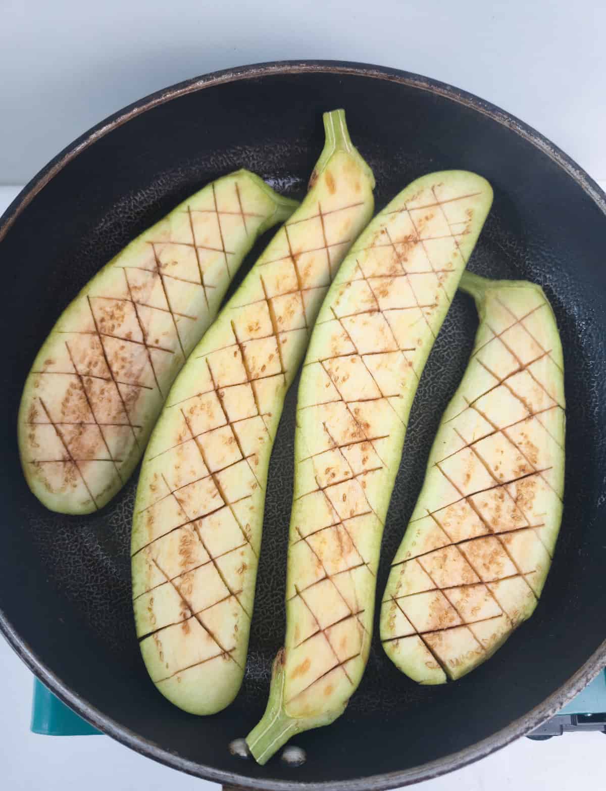 Crosshatched eggplant halves arranged cut side up in a skillet before cooking.
