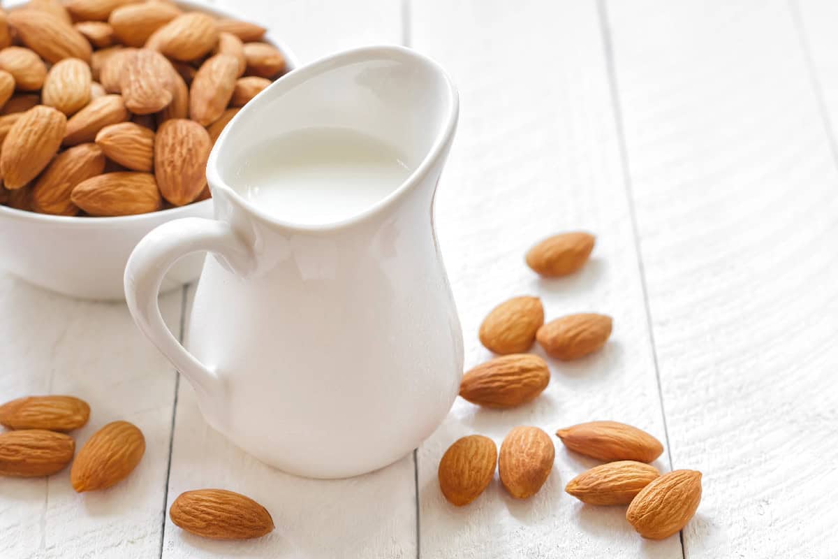 Creamy almond milk in a white pitcher surrounded by whole almonds on a white wooden table.