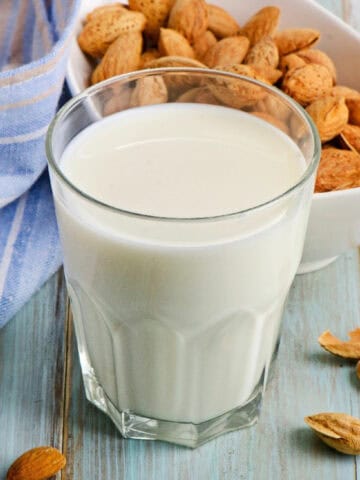 Glass of almond milk with almonds and green leaves on a blue wooden surface.