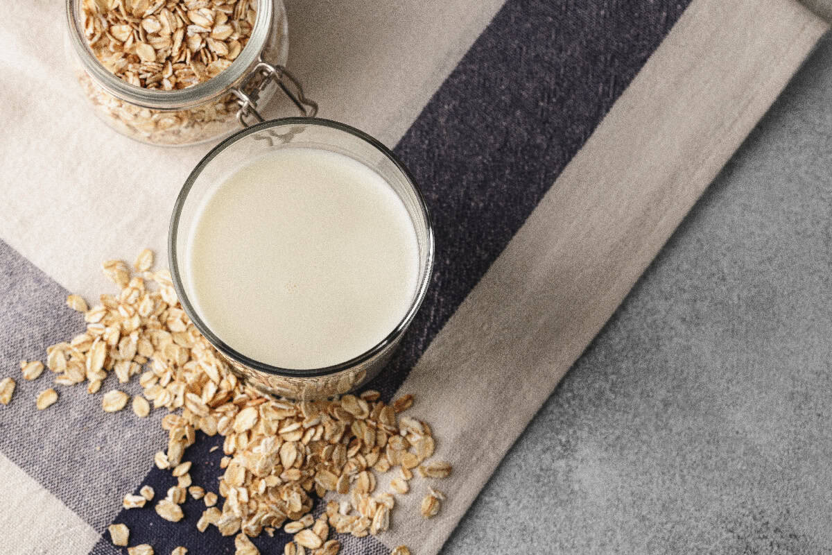 Glass of oat milk surrounded by rolled oats on a striped cloth with a jar of oats in the background.