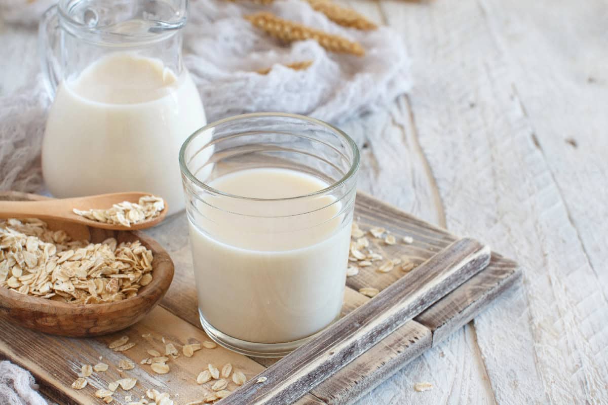 Homemade oat milk in a glass with rolled oats and a small milk pitcher on a wooden tray.