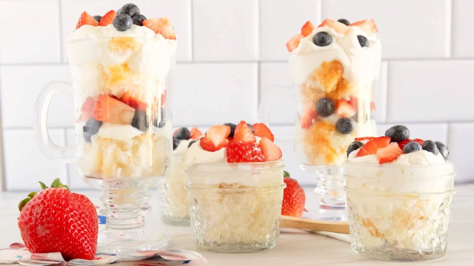 Red, white, and blue trifle desserts made with angel food cake, vanilla pudding, whipped topping, strawberries, and blueberries displayed in individual glass serving cups.