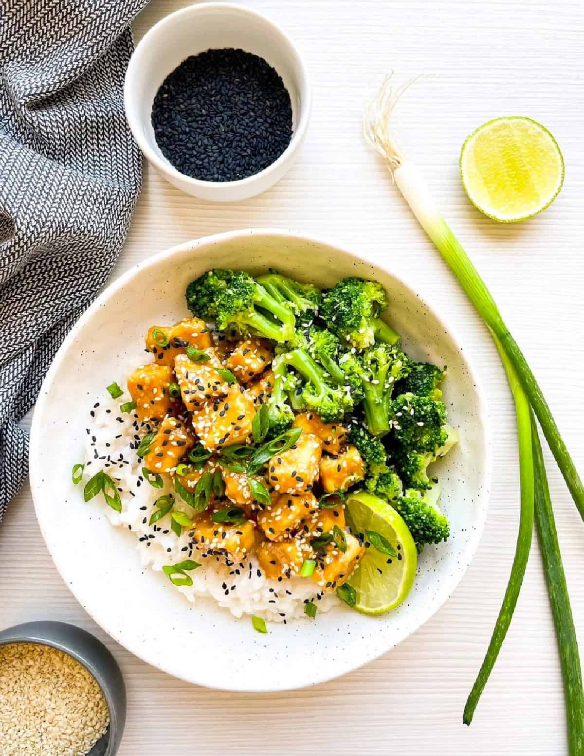Sesame tofu with broccoli served over steamed rice with scallions, sesame seeds, and a lime wedge.