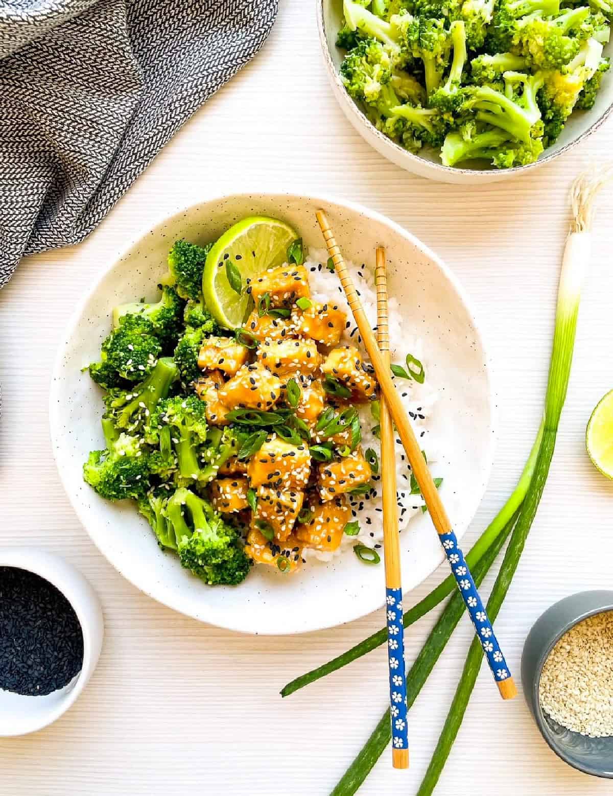 Healthy sesame tofu with broccoli over steamed rice garnished with scallions, sesame seeds, and lime.