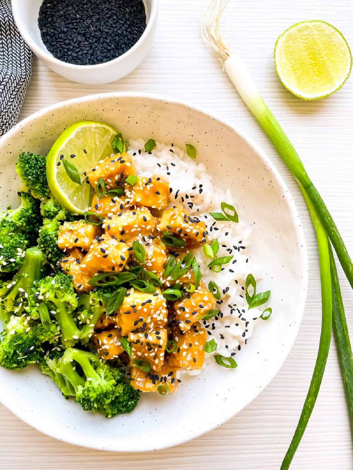 Close-up of sesame tofu, broccoli, and white rice topped with sesame seeds and sliced scallions.