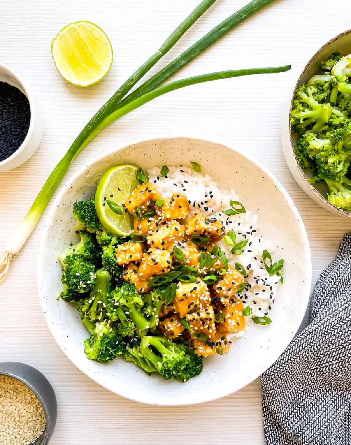 Overhead view of sesame tofu with broccoli served over rice in a bowl with fresh lime.