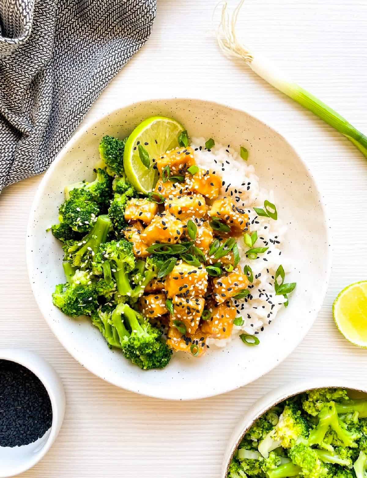 Healthy sesame tofu rice bowl with broccoli, lime wedges, and sesame seed garnish.