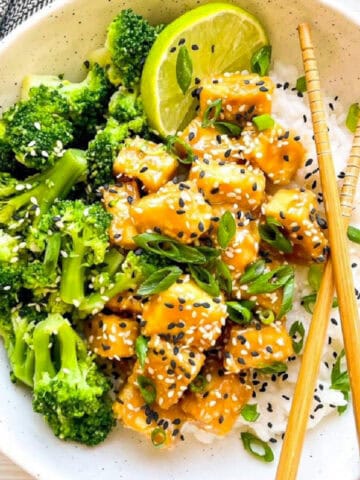 Sesame tofu with broccoli served over steamed rice, topped with sesame seeds, sliced scallions, and a fresh lime wedge.