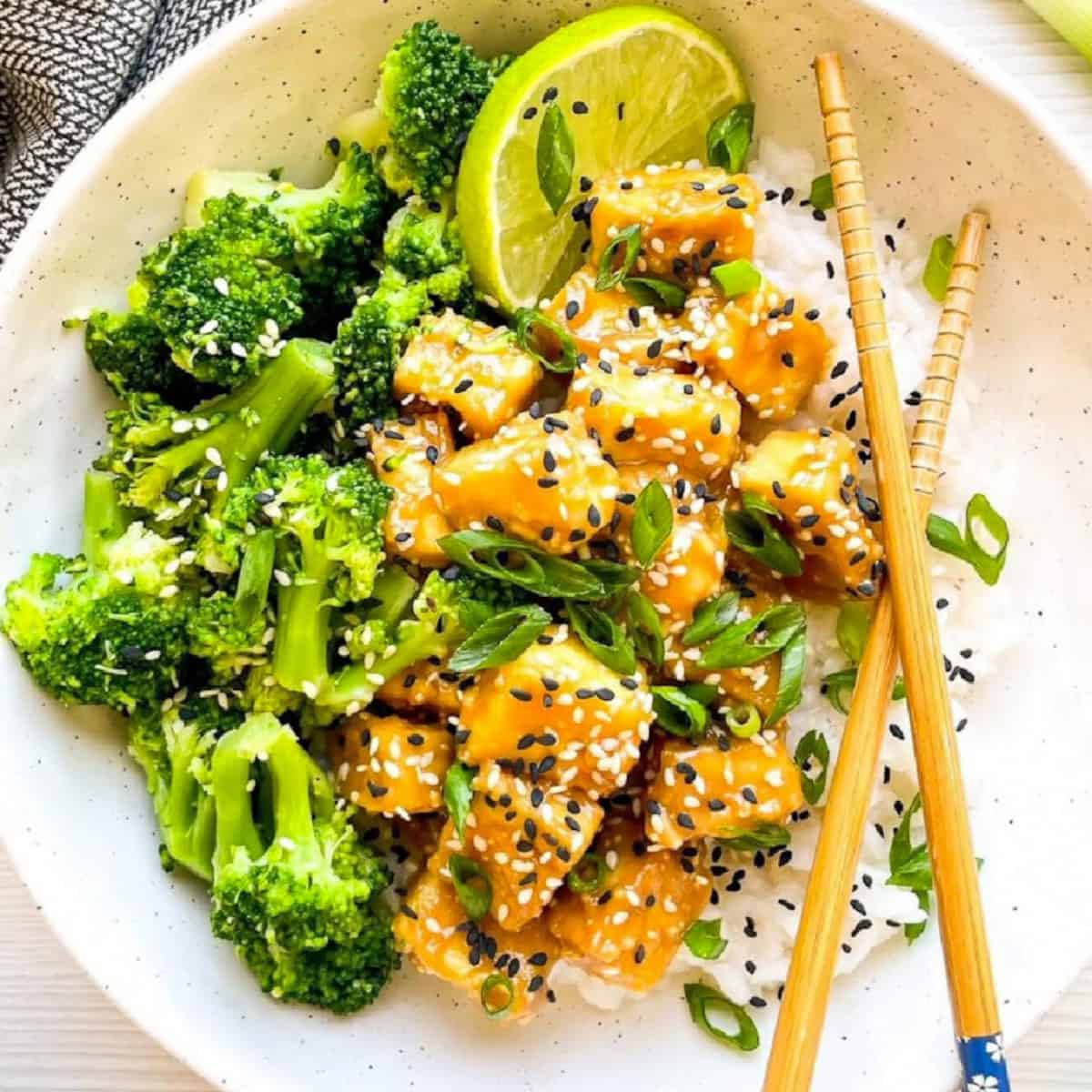 Sesame tofu with broccoli served over steamed rice, topped with sesame seeds, sliced scallions, and a fresh lime wedge.