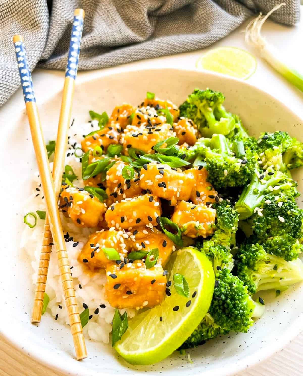 Sesame tofu rice bowl with broccoli, chopsticks, lime, and sesame seed garnish.
