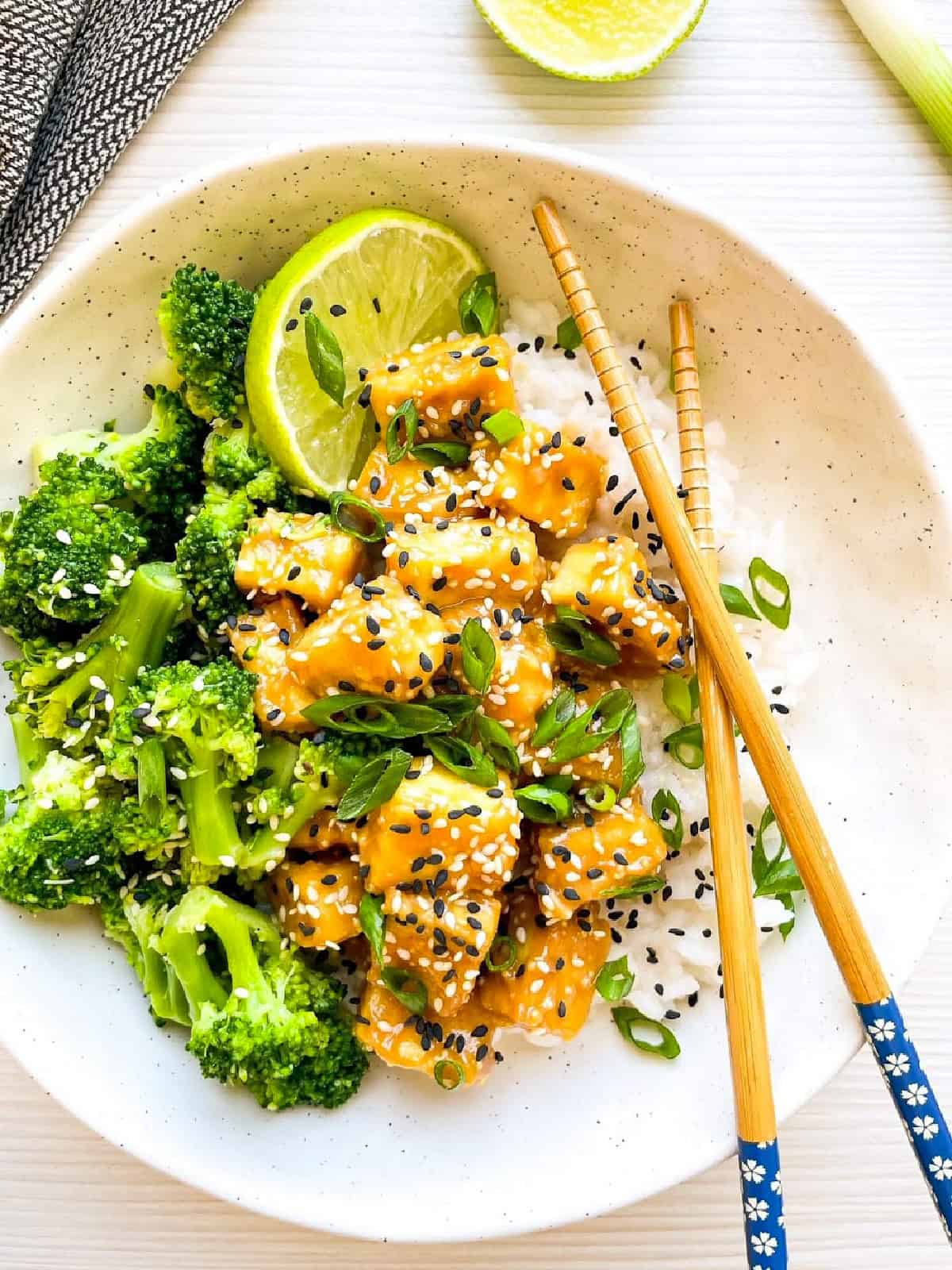 Overhead view of sesame tofu with broccoli served over rice with chopsticks and lime.