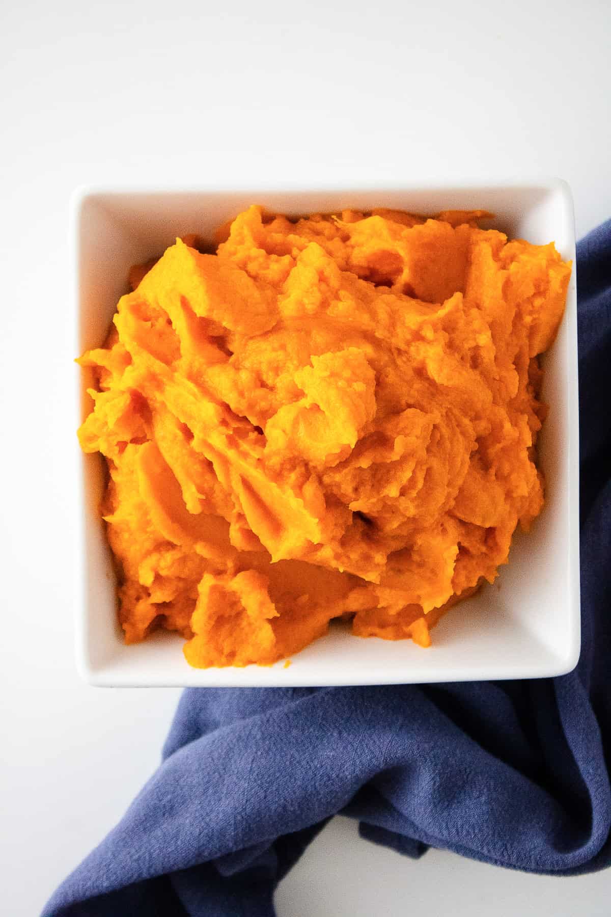 Overhead view of homemade sweet potato puree in a white square bowl with a blue napkin.