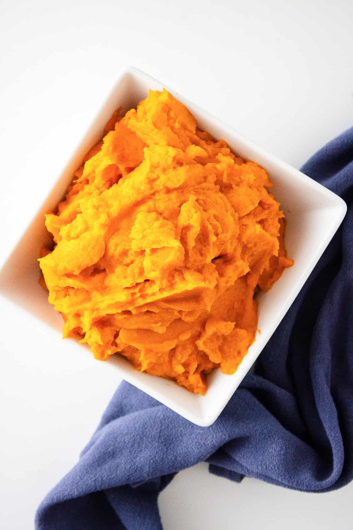 Overhead shot of sweet potato puree in a white bowl with a blue cloth napkin beside it.