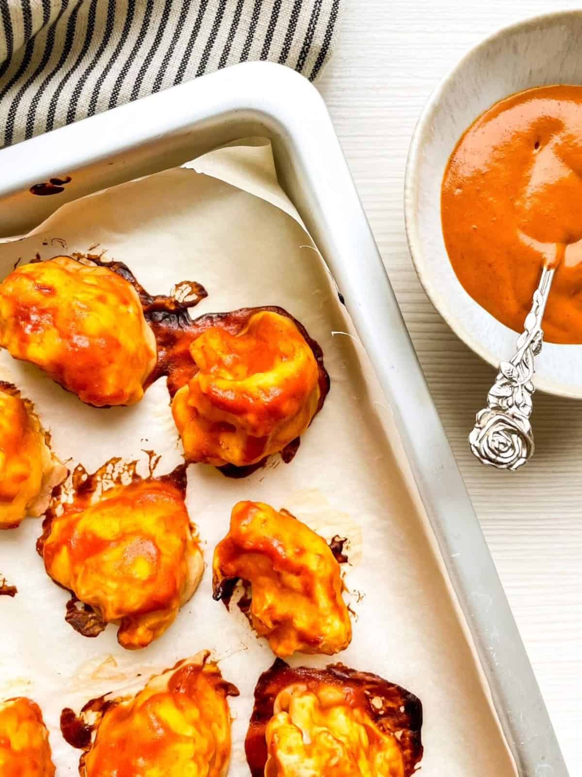 Buffalo sauce-coated cauliflower wings on a parchment-lined baking sheet next to a bowl of buffalo sauce.
