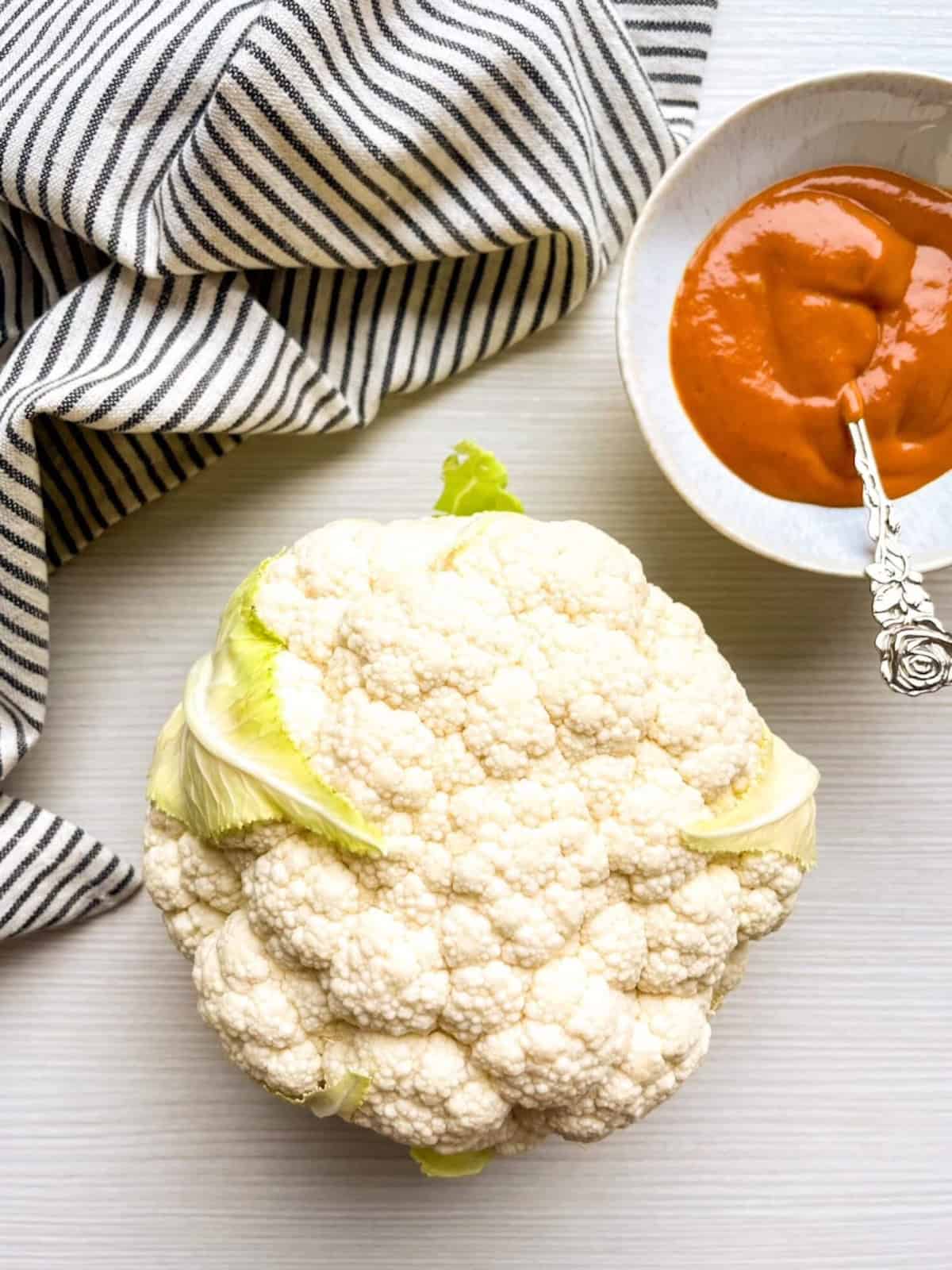 Whole cauliflower head on a light tabletop next to a bowl of buffalo sauce and a striped kitchen towel.