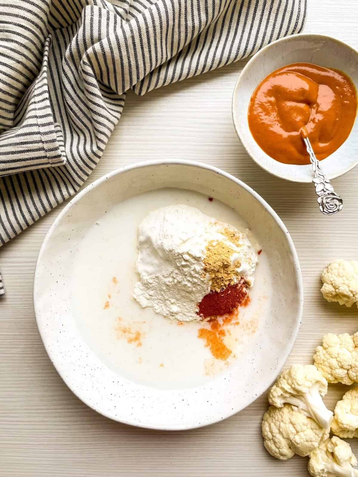 Flour, paprika, garlic powder, and plant-based milk in a mixing bowl with cauliflower florets nearby.