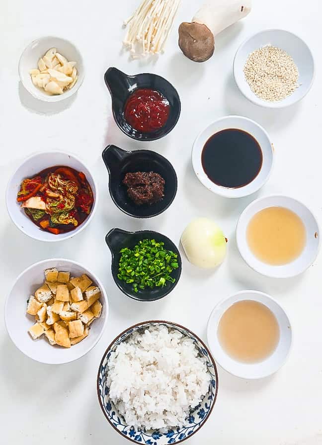 Ingredients for vegan kimchi fried rice, including cooked rice, kimchi, fried tofu puffs, mushrooms, garlic, onion, green onions, soy sauce, sesame seeds, and seasonings arranged on a white surface.