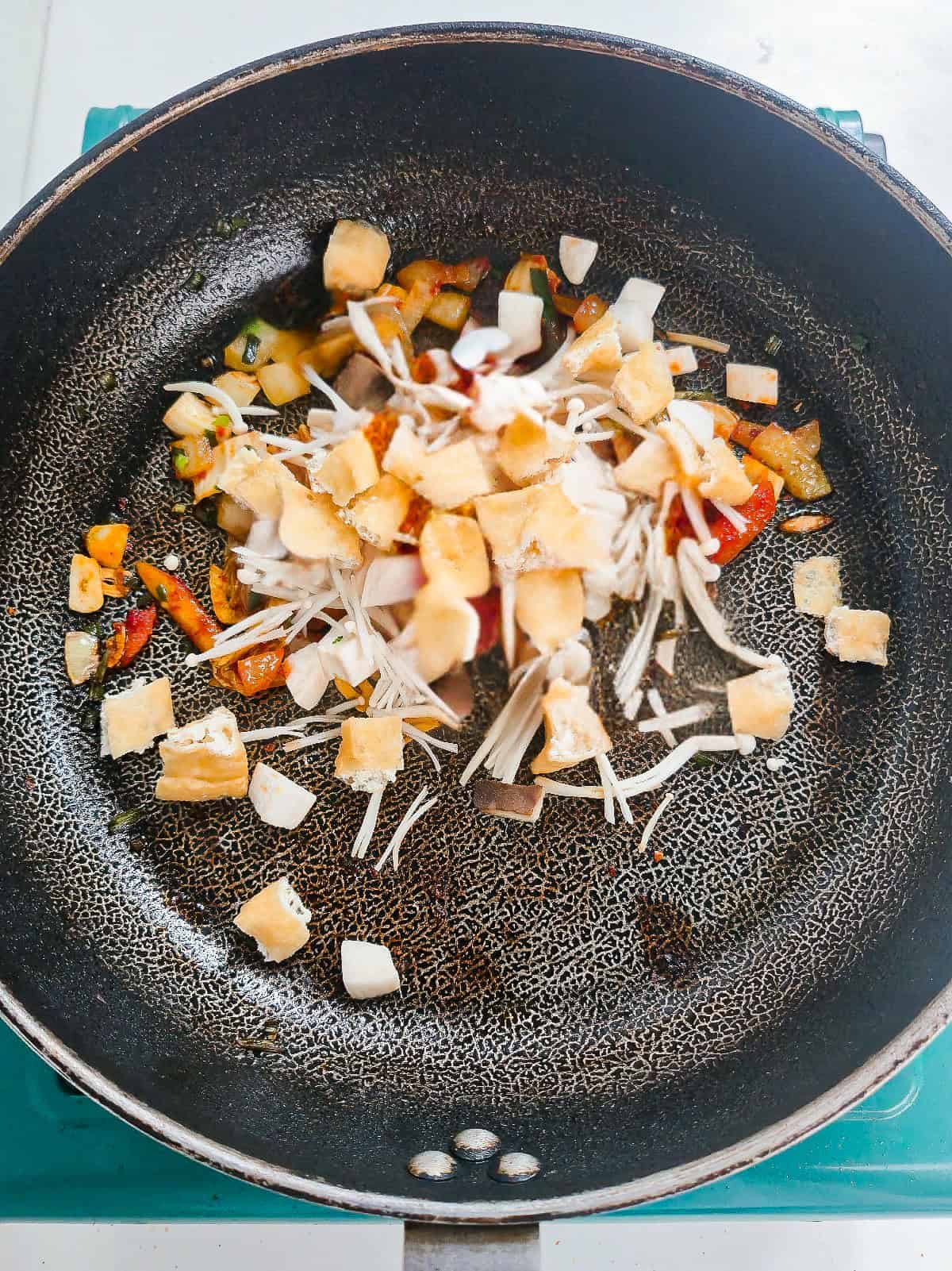 Fried tofu puffs and mushrooms added to kimchi and aromatics in a skillet.