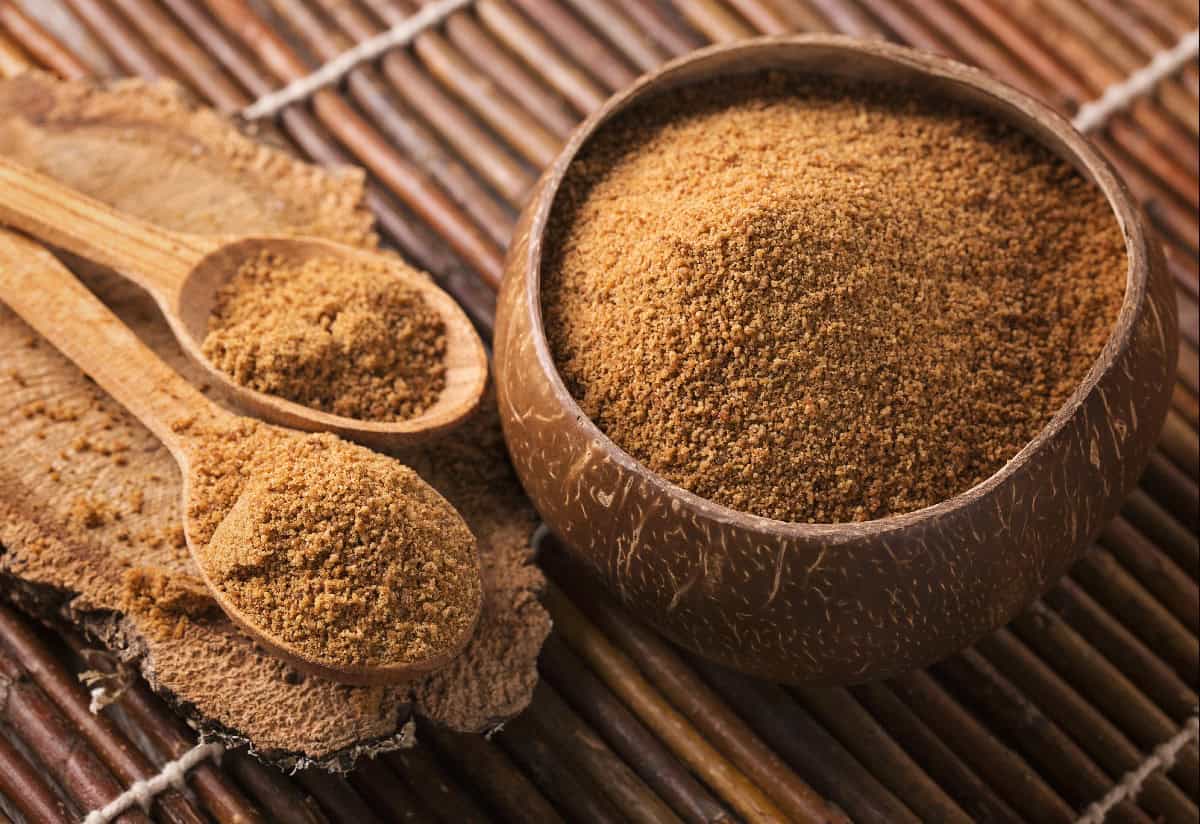 Coconut sugar in wooden spoons and a coconut shell bowl on a bamboo mat.