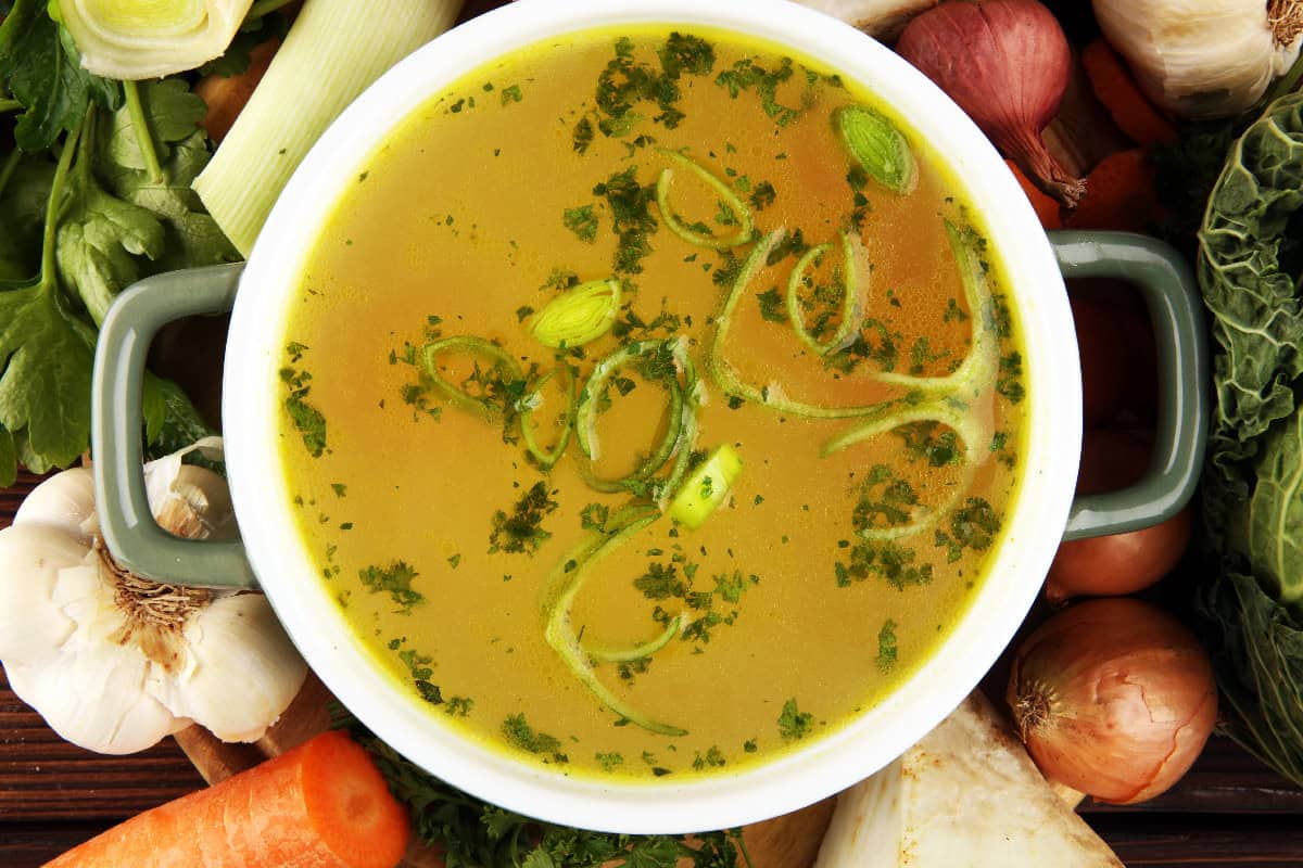 Homemade vegetable broth garnished with herbs and surrounded by fresh vegetables.