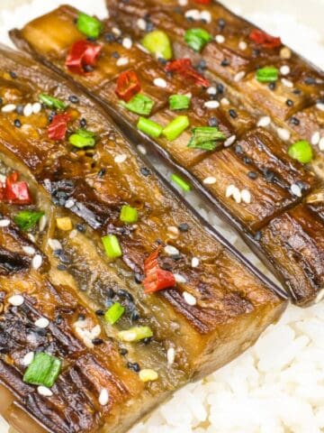 Close-up of vegan unagi-style eggplant served over steamed white rice, topped with sesame seeds, chopped green onions, and red chili pieces.