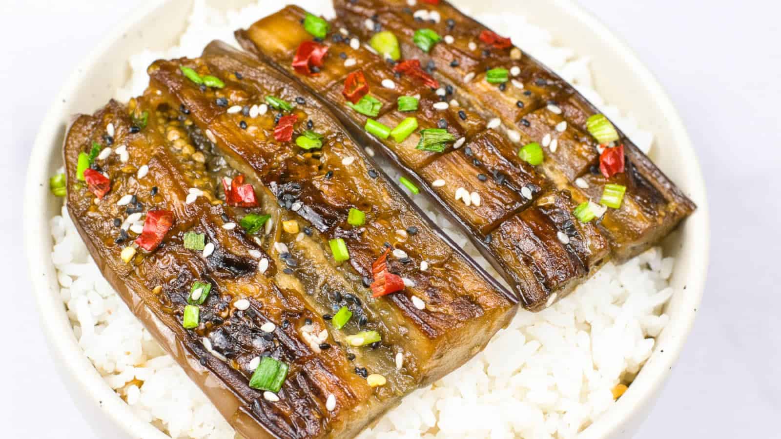 Close-up of vegan unagi-style eggplant served over steamed white rice, topped with sesame seeds, chopped green onions, and red chili pieces.