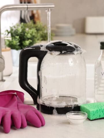 Electric kettle with soap suds, vinegar, sponge, and cleaning supplies on a kitchen counter.