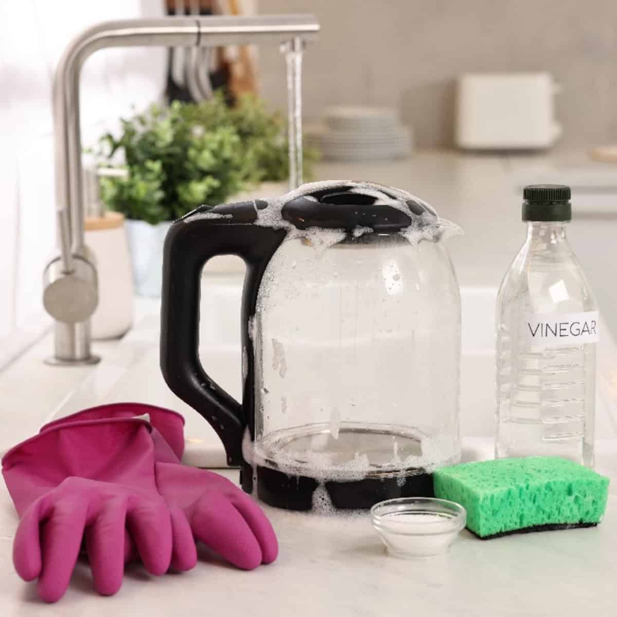 Electric kettle with soap suds, vinegar, sponge, and cleaning supplies on a kitchen counter.